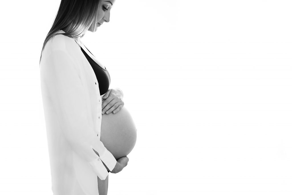 Healthy Pregnancy - Pregnant woman looking down on her belly wearing a white shirt and resting her hands on her belly