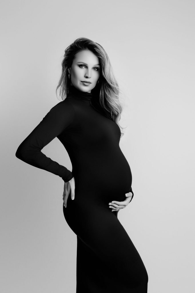 A pregnant woman in a long-sleeved, fitted dress poses in profile with one hand on her hip and the other on her belly against a plain background, perfect for newborn photography Vancouver sessions.