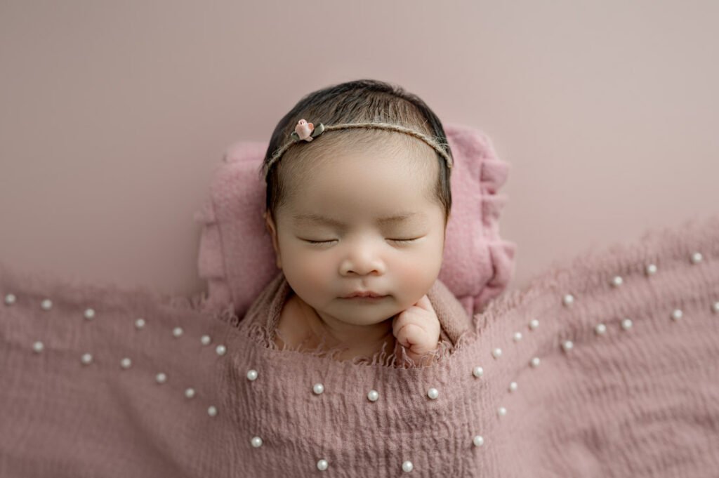 A newborn baby sleeps on a pink blanket with a matching headband and a textured, pearl-decorated covering, capturing the sweetness of Newborn Photography Vancouver.