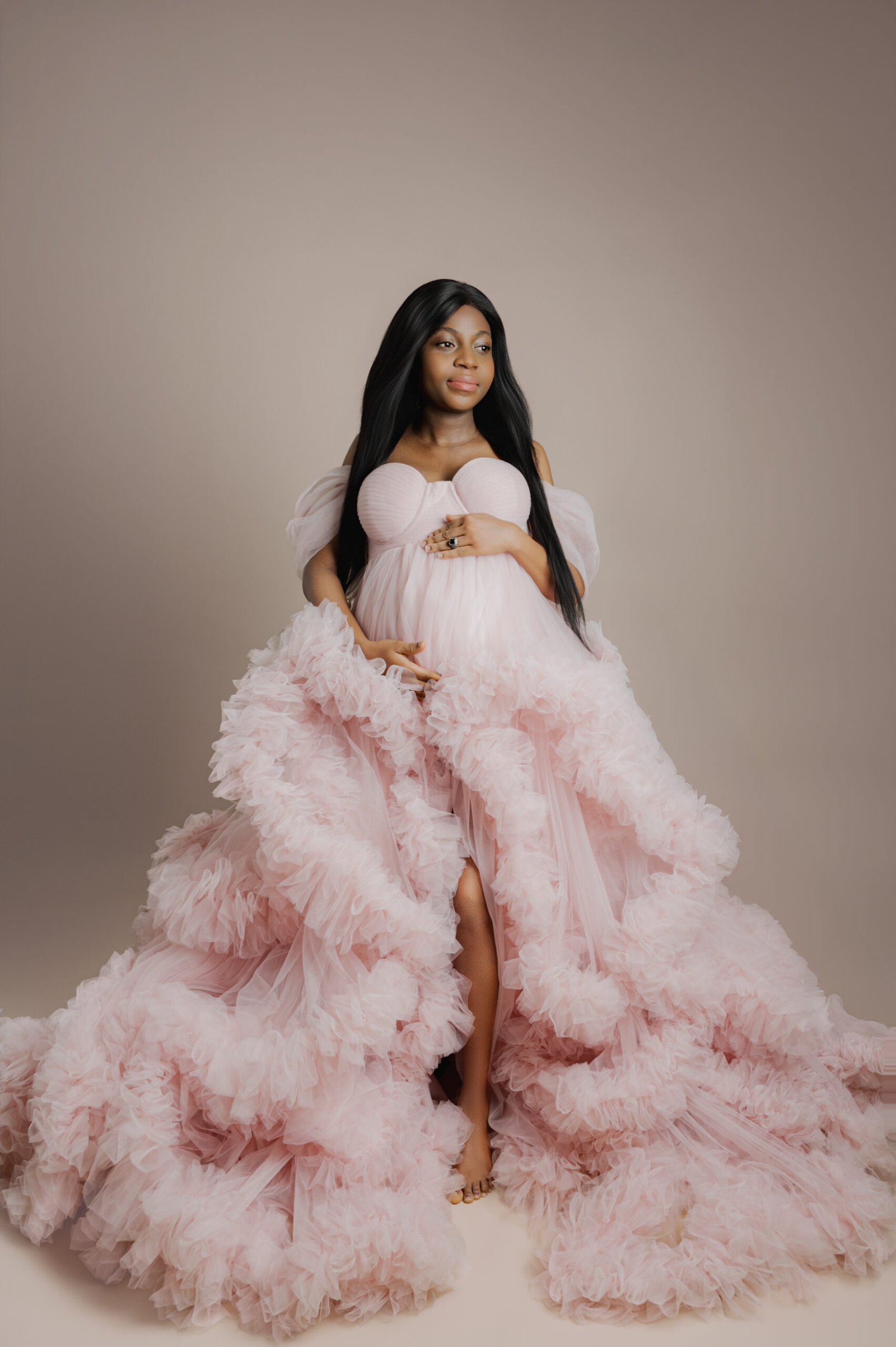 A woman in a pink, ruffled gown poses against a neutral background, holding her pregnant belly and looking slightly to the side.