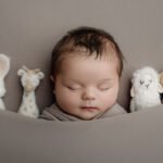 A baby is sleeping on a gray surface, wrapped in a blanket, with four plush toys—a rabbit, giraffe, lamb, and bear—lined up beside them. Capture sweet moments like these with Newborn Photography Vancouver.