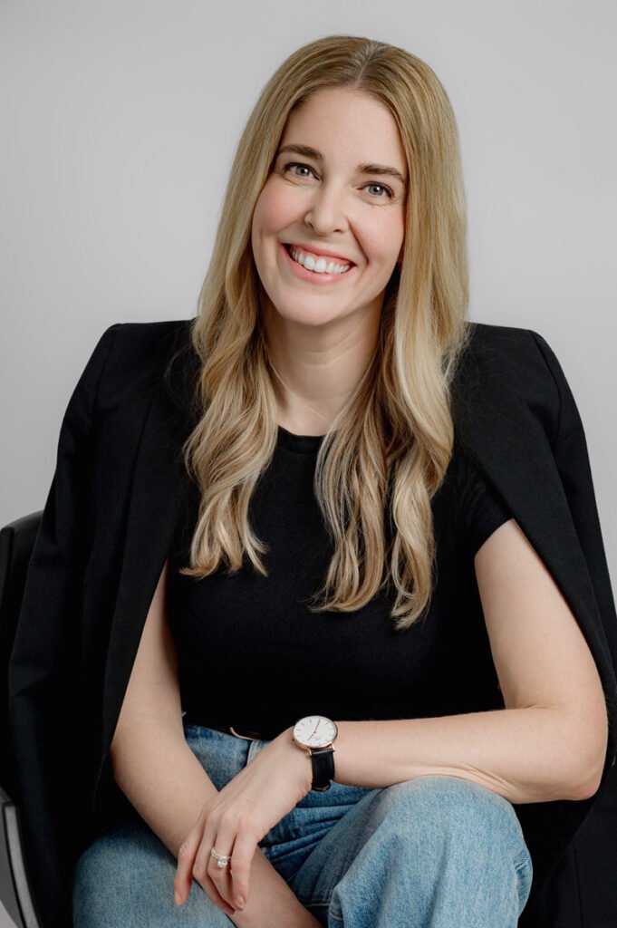 A woman with long blonde hair, wearing a black top, blazer, and jeans, sits on a chair, smiling at the camera.