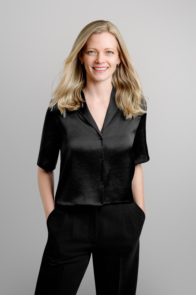 Woman with blonde hair wearing a black short-sleeve blouse and black pants, standing with hands in pockets against a plain light gray background—perfect for womens headshots.