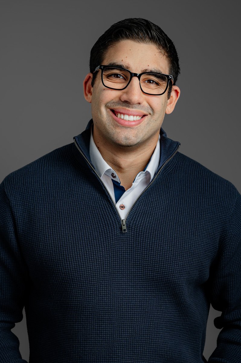 A man wearing glasses and a dark blue zip-up sweater over a collared shirt smiles at the camera against a plain gray background.