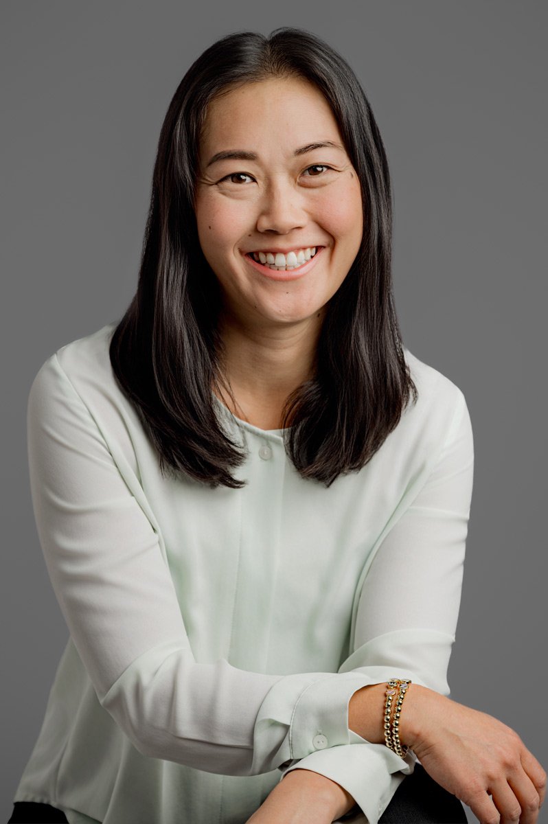 A woman with long dark hair, wearing a light blouse and bracelets, sits and smiles at the camera against a plain gray background.