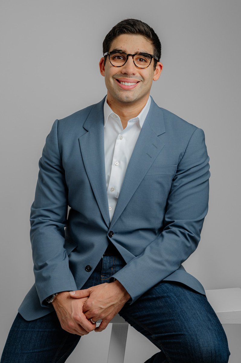A man wearing glasses, a blue blazer, white shirt, and jeans sits on a white stool against a plain gray background, smiling at the camera.