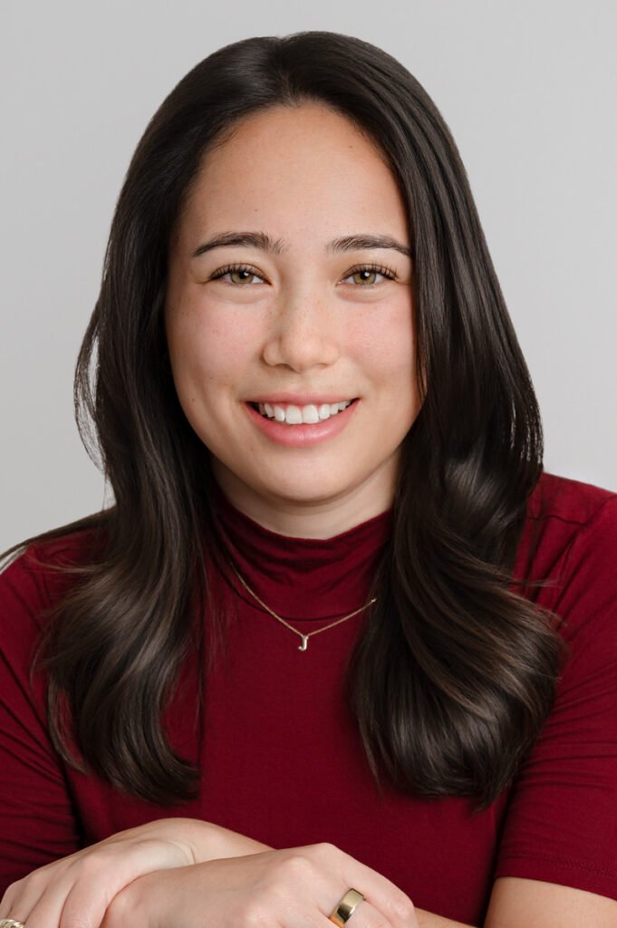 A woman with long dark hair and a maroon top smiles at the camera in this headshot, resting her hands on a surface. She wears a delicate necklace and a ring. The background is light gray.