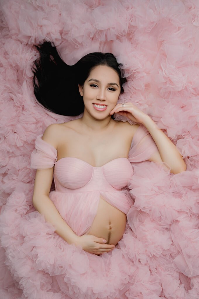 A pregnant woman in a pink tulle dress lies on a matching pink background, smiling and holding her belly—capturing the magic of motherhood with T Haller Photography, specialists in newborn photography Vancouver.