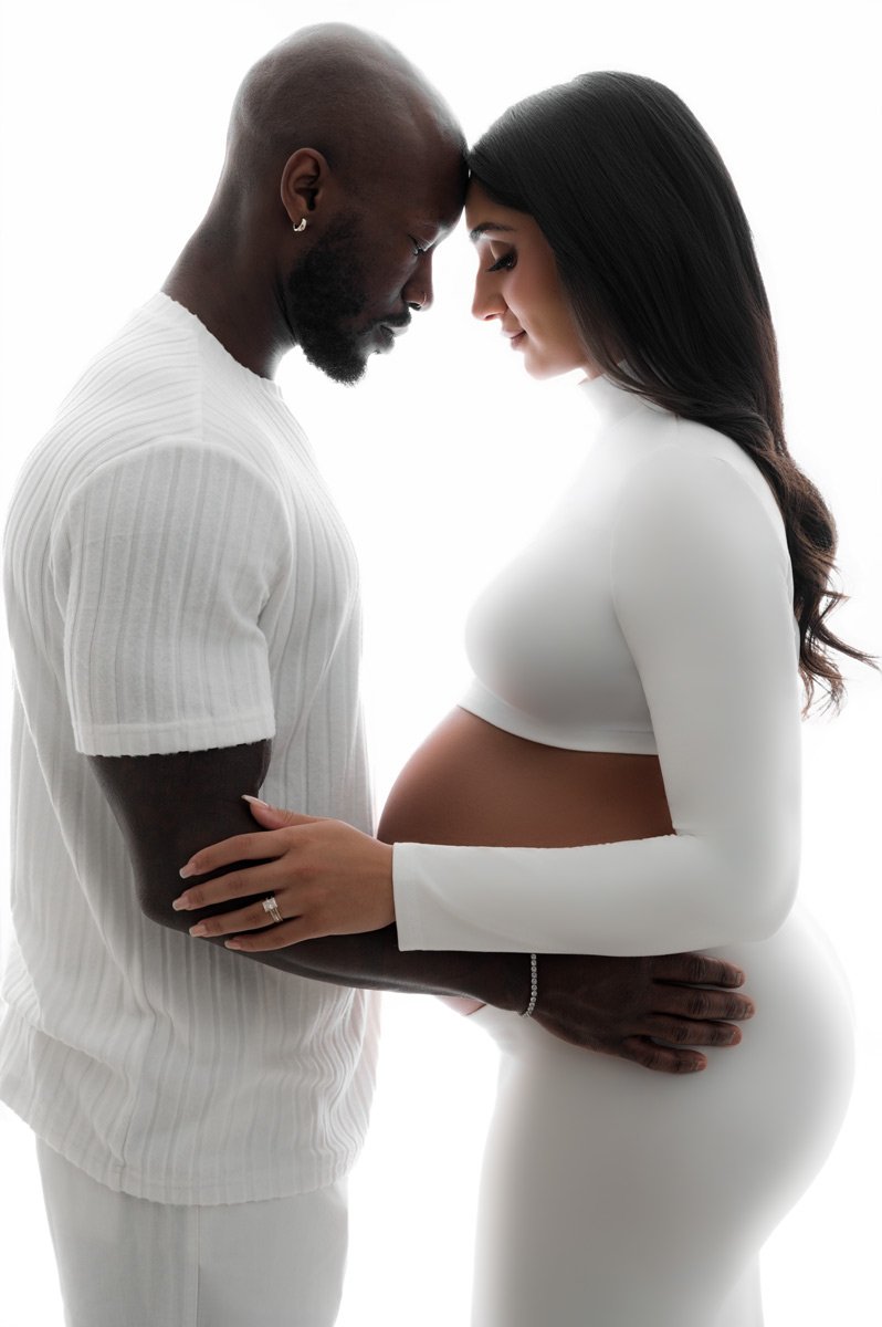 A man and a pregnant woman stand face to face, gently touching foreheads and holding her belly, both dressed in white against a white background—a serene moment captured by T Haller Photography, specialists in newborn photography Vancouver.
