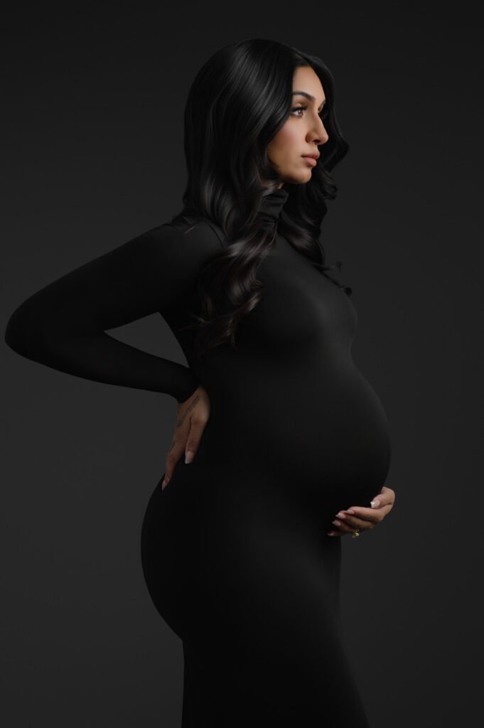 Pregnant woman in a fitted black dress stands in profile, one hand on her lower back and the other on her stomach, against a dark background—capturing a timeless moment by T Haller Photography, specialists in newborn photography Vancouver.