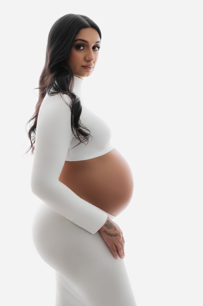A pregnant woman in a white fitted outfit poses in profile against a white background, her hand gently resting on her belly—a serene moment captured by T Haller Photography, specialists in newborn photography Vancouver.
