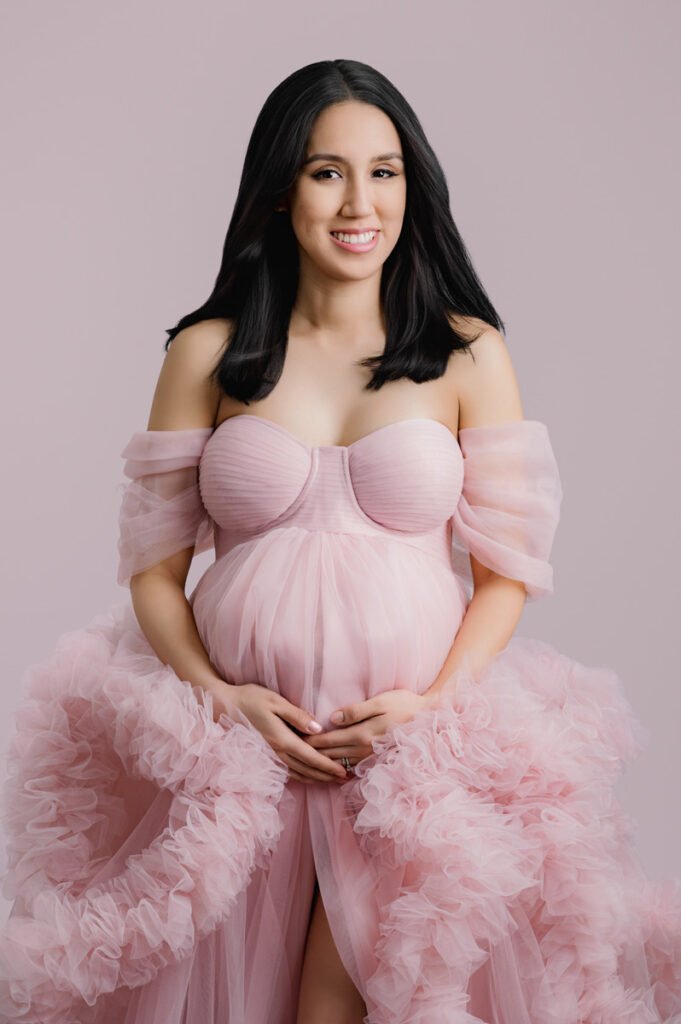 A pregnant woman in a pink off-shoulder tulle gown stands against a light pink background, smiling and holding her hands on her belly. Captured by T Haller Photography, specialists in newborn photography Vancouver moms love.