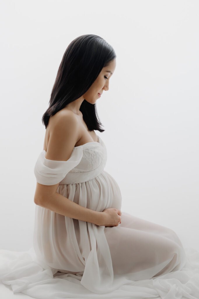 A pregnant woman in a sheer white dress sits on the floor in profile against a plain white background, gently holding her belly—a serene moment beautifully captured by T Haller Photography, specialists in newborn photography Vancouver.