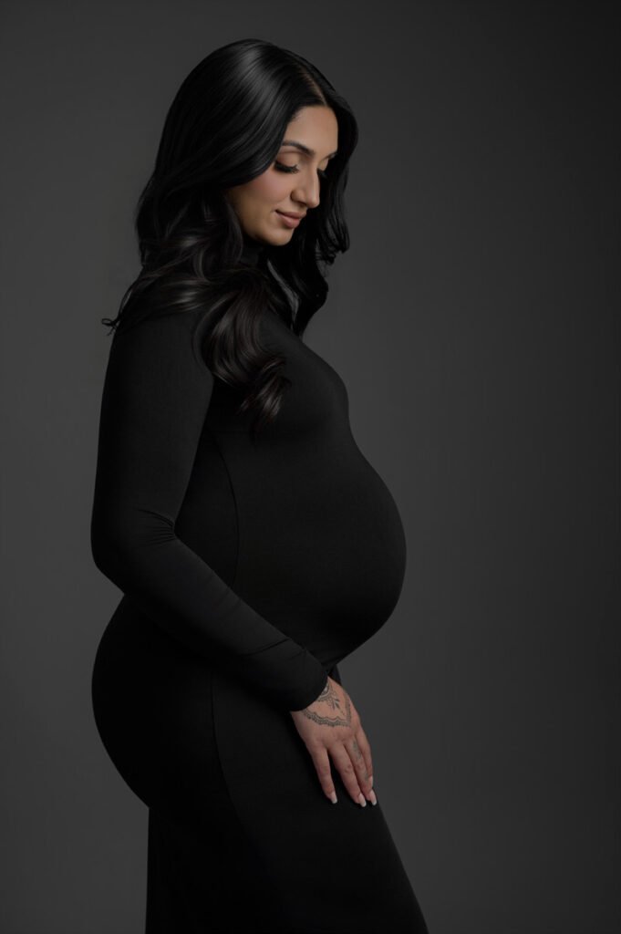 Pregnant woman in a black dress standing in profile against a dark background, gently resting her hand on her belly. Captured by T Haller Photography, specialists in maternity and newborn photography Vancouver.