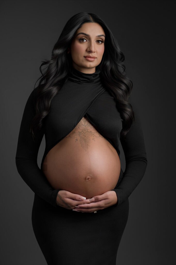 A pregnant woman in a long-sleeved black dress poses with her hands on her exposed belly, against a dark background—an elegant maternity portrait by T Haller Photography, specialists in newborn photography Vancouver.