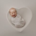 A newborn baby wrapped in a white blanket is lying in a white, heart-shaped bowl with a neutral background, perfectly styled for a newborn photographer’s portfolio.
