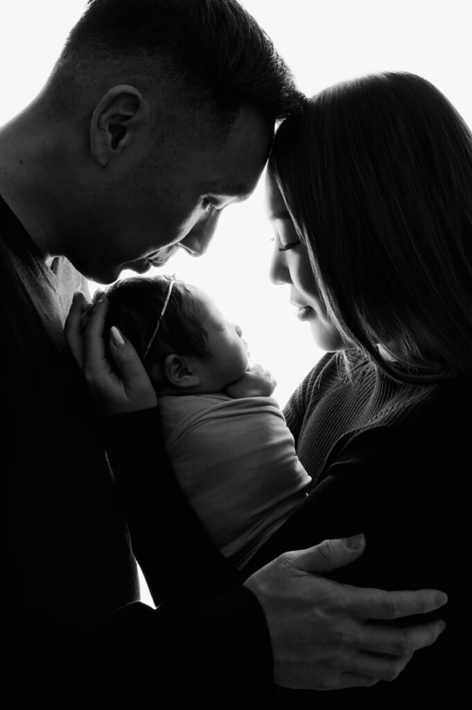 Black and white photo by a newborn photographer: two adults gently touch foreheads while holding a baby between them, facing each other in a close, intimate pose.