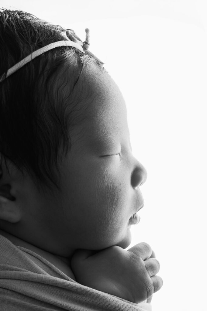 Black and white side profile of a sleeping newborn baby with eyes closed, headband on, and hands resting under the chin against a white background—captured by a skilled newborn photographer.
