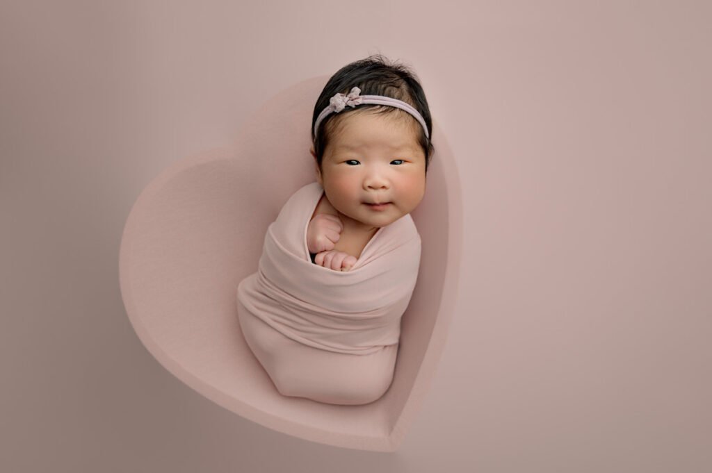 A newborn photographer captures a baby wrapped in a pink blanket, lying in a heart-shaped cushion, with a pink headband, against a pale pink background.
