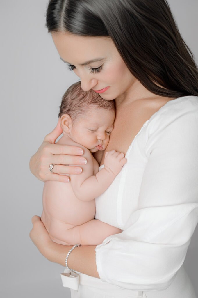 A newborn photographer captures a tender moment as a woman in a white blouse gently holds a sleeping newborn baby against her chest, both facing forward against a plain background.