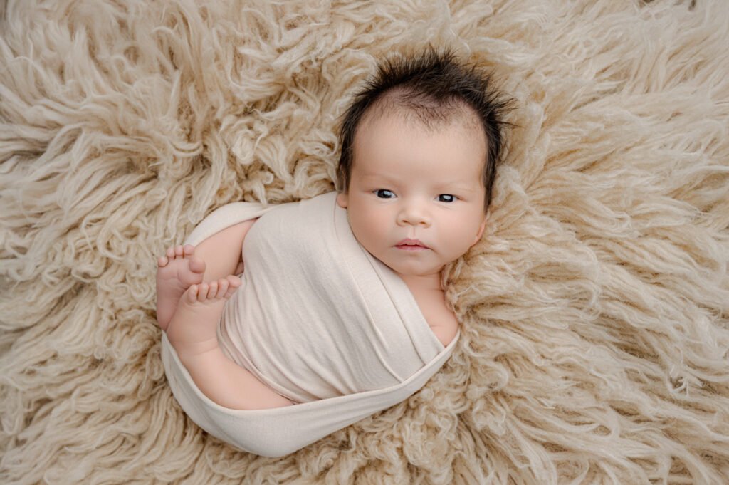 A baby wrapped in a cream-colored blanket lies on a soft, fluffy beige rug, looking up with a calm expression—perfect inspiration for any newborn photographer.