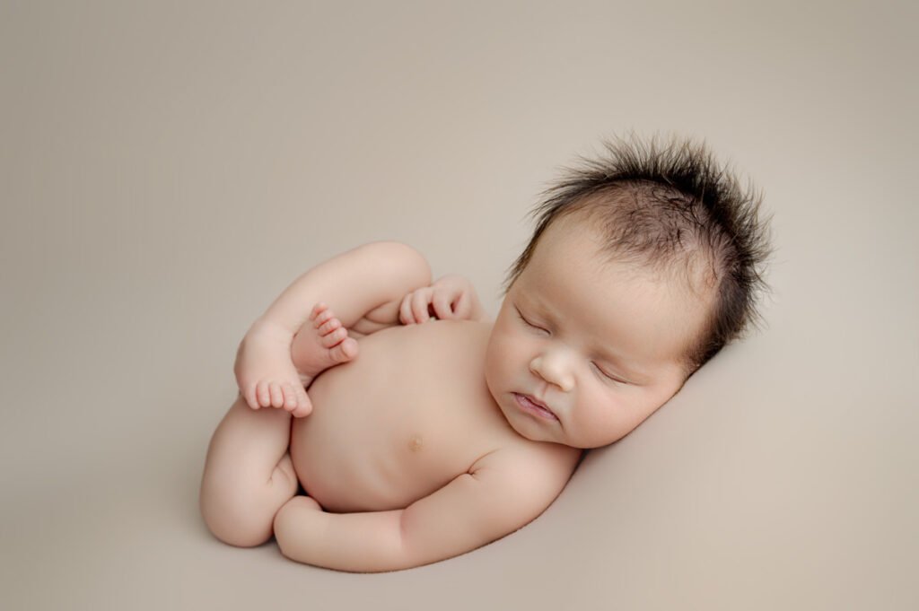 A newborn photographer captures a baby with dark hair lying on a neutral beige background, peacefully sleeping on their back with hands and feet relaxed.
