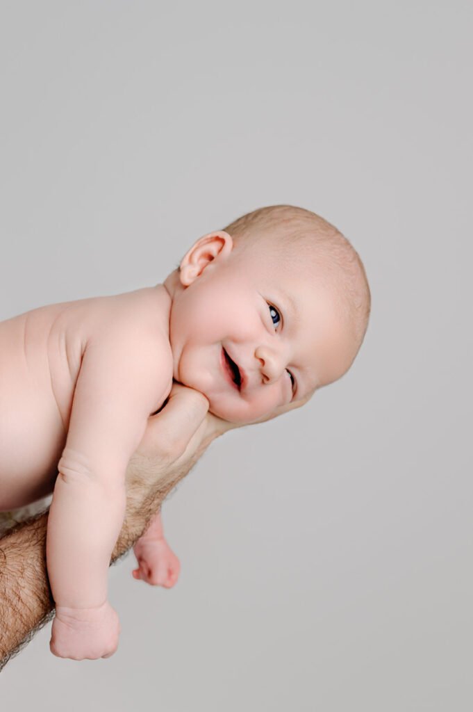 An adult hand holds a smiling baby, gently posed on their stomach against a plain light gray background—a perfect moment for a newborn photographer to capture.