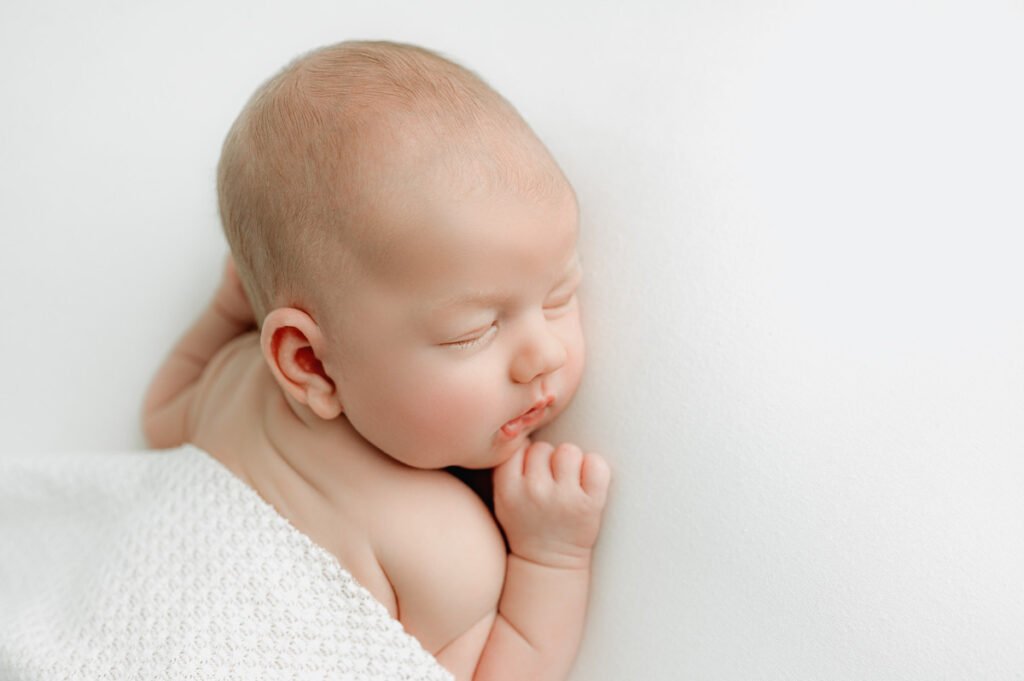 Newborn baby sleeping on a white surface, partially covered with a white textured blanket, with one hand near their face—an ideal scene for a newborn photographer to capture precious early moments.