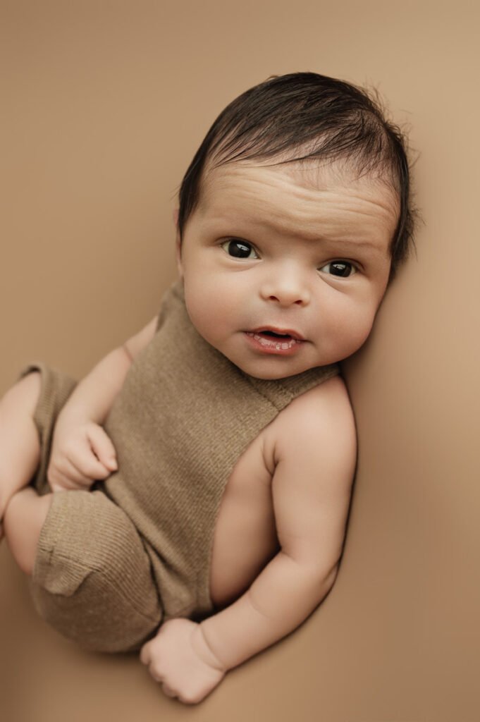 A baby with dark hair and brown eyes, captured by a newborn photographer, sits in a brown sleeveless outfit on a matching background, looking up at the camera.