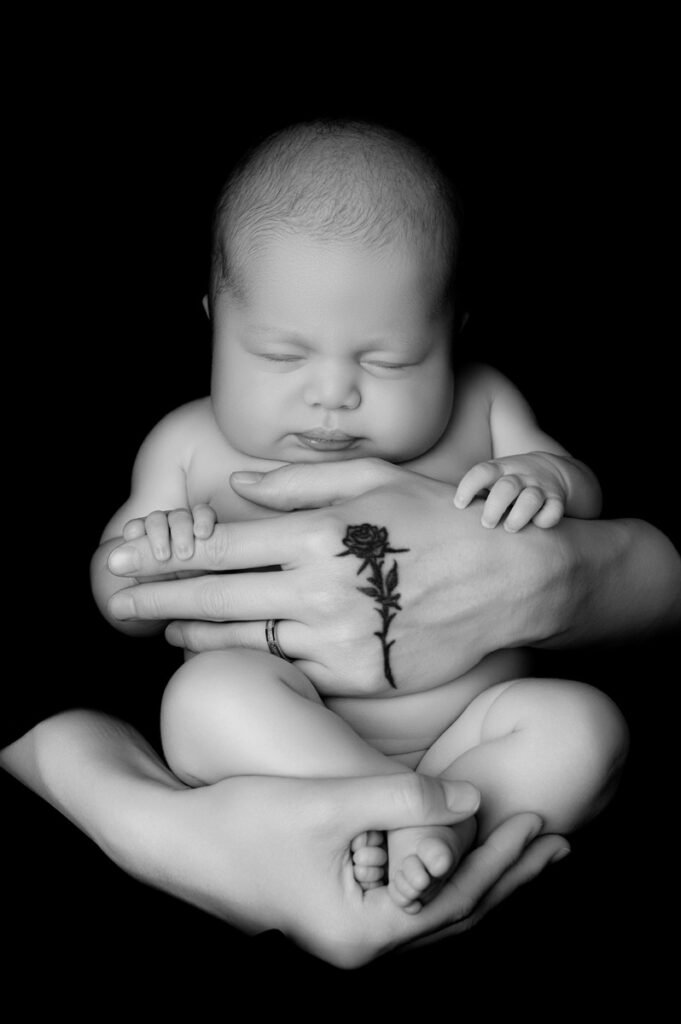 A newborn photographer captures a baby cradled by adult hands, one adorned with a rose tattoo and ring, set against a black background.