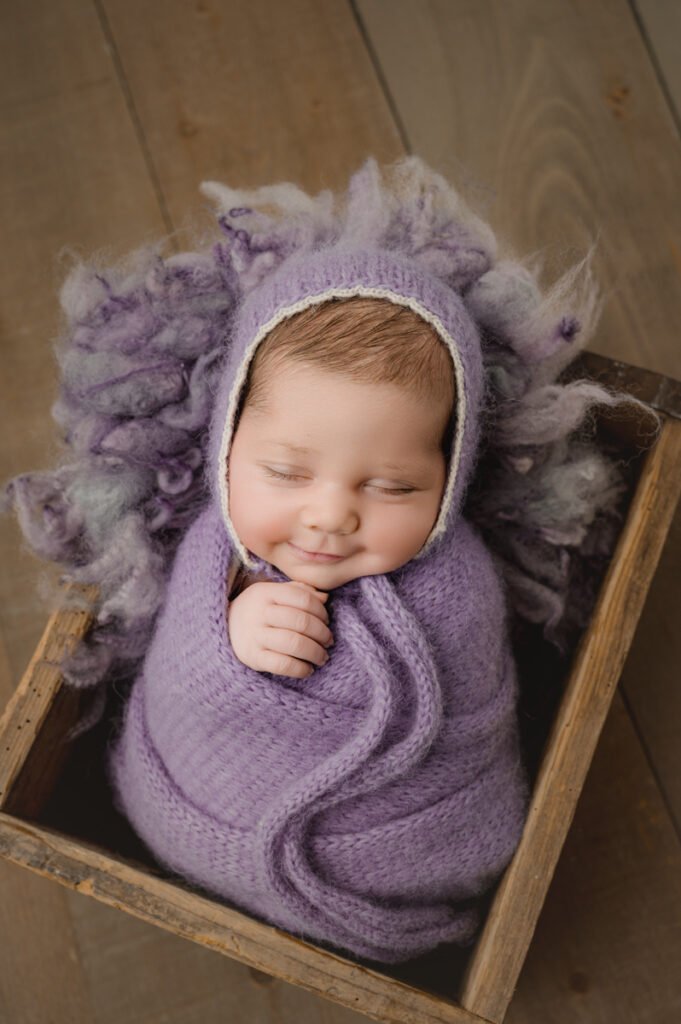 A sleeping baby, wrapped in a purple knit blanket and bonnet, lies in a wooden box on a wooden floor—an adorable moment perfectly captured by a skilled newborn photographer.