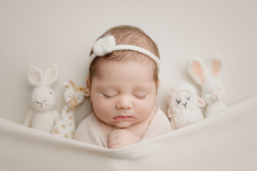 A sleeping baby with a white headband is tucked under a blanket, surrounded by four stuffed animal toys on a light background—an adorable scene captured perfectly by a skilled newborn photographer.