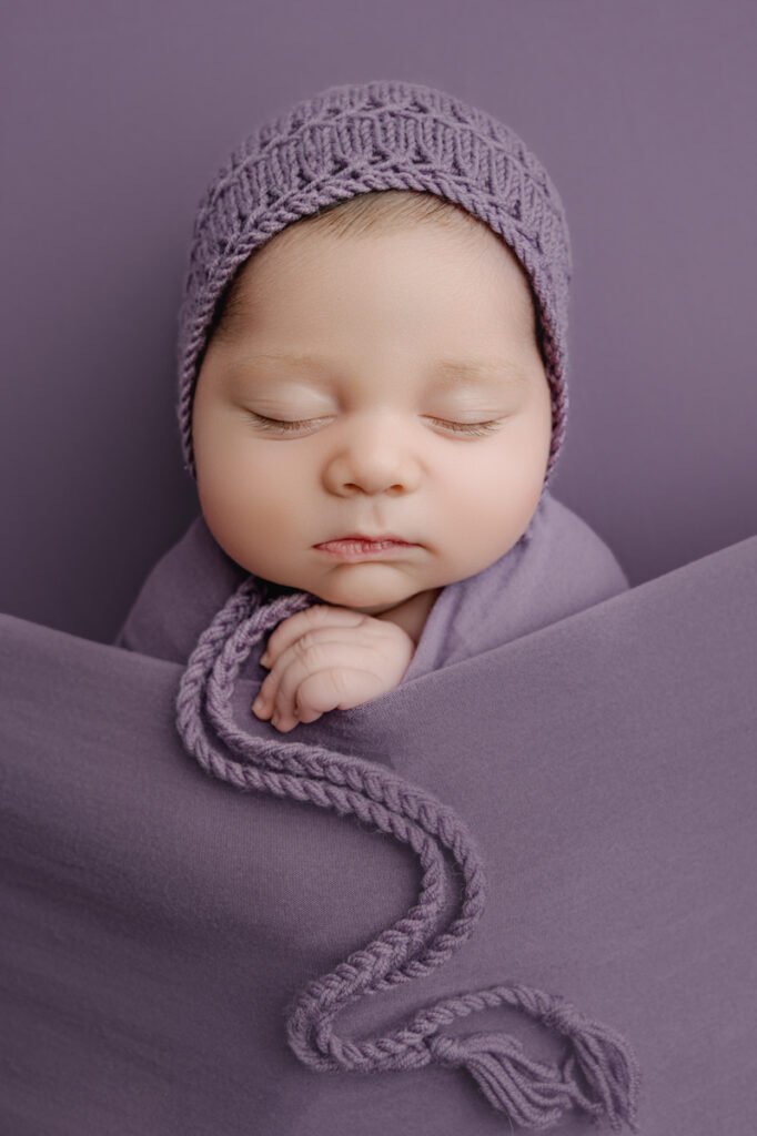 Newborn photographer captures a peaceful baby sleeping, wrapped in a purple blanket and wearing a matching knitted hat, against a soft purple background, for West Vancouver families