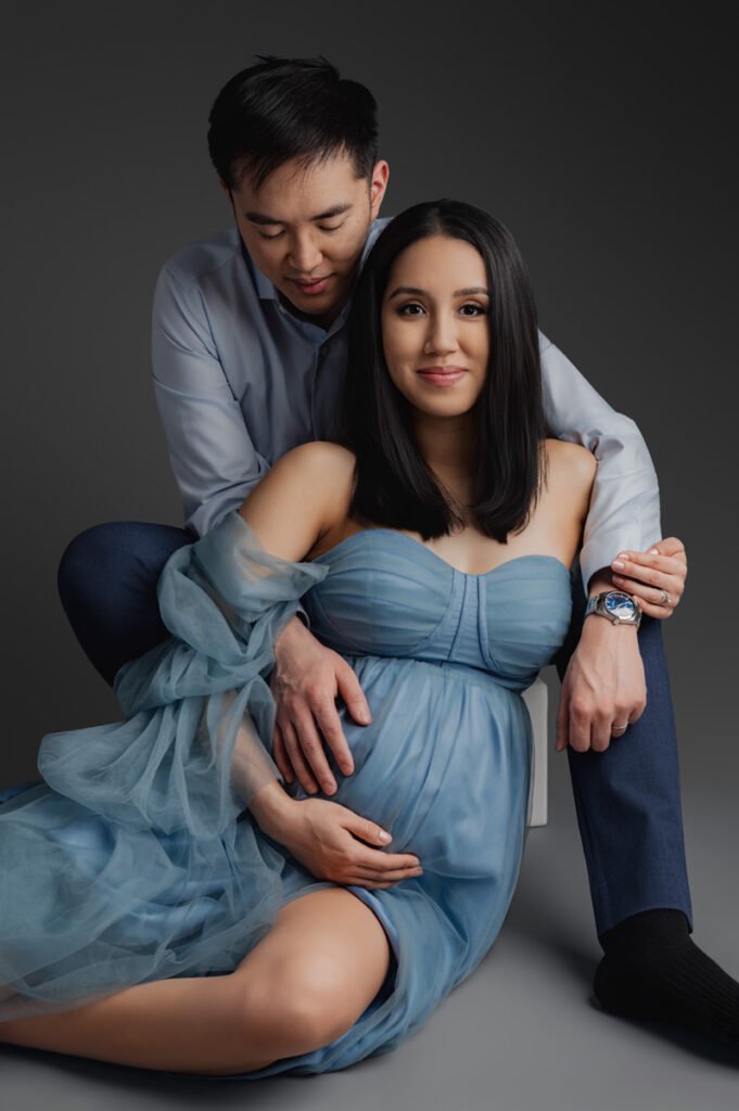 A couple poses together for T Haller Photography; the woman, in a blue gown, is pregnant while the man sits behind her with a hand on her belly. They both look at the camera, capturing a special moment before their newborn photography Vancouver session.