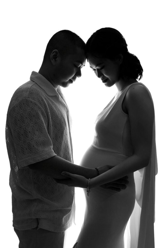A man and a pregnant woman stand facing each other in profile, touching foreheads and holding the woman's belly against a white background—a tender moment captured in beautiful maternity photography.
