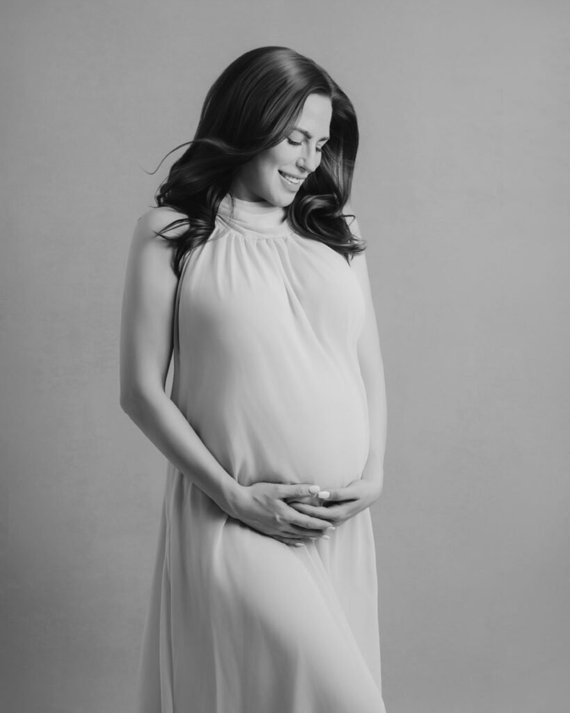 A woman in a long, sleeveless dress stands holding her pregnant belly with both hands, smiling and looking down. This elegant black and white image beautifully captures the essence of maternity photography.