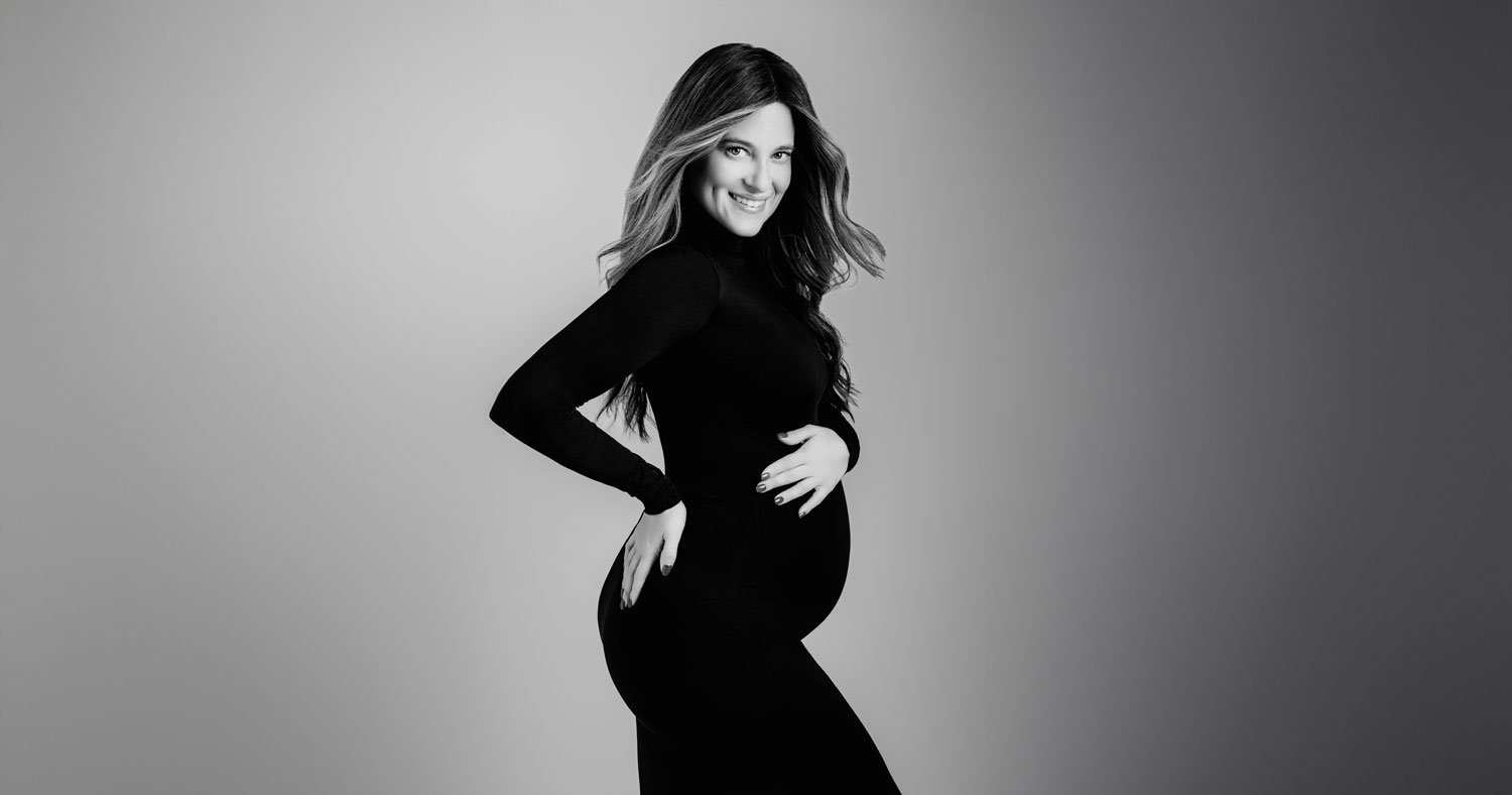 A pregnant woman in a long-sleeved, form-fitting black dress poses sideways, holding her belly and smiling, beautifully capturing the essence of maternity photography against a neutral background.