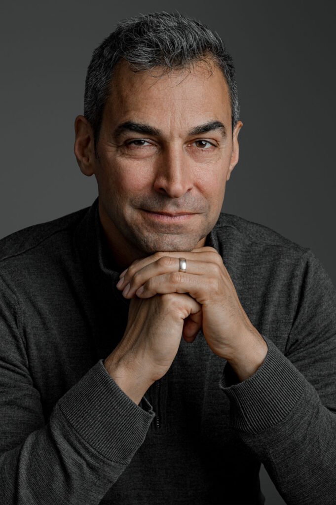 A man with short gray hair and a trimmed beard sits with hands clasped, wearing a dark zip-up sweater and a wedding ring, against a plain gray background—an ideal setting for professional headshots.