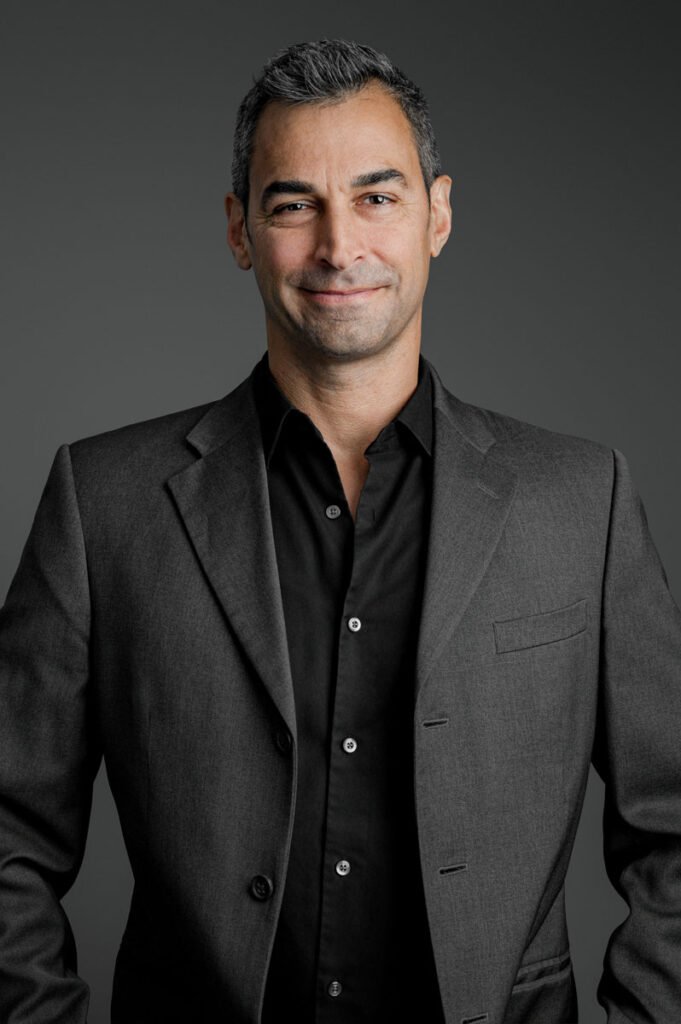 A man with short dark hair wearing a charcoal suit jacket and black shirt stands against a plain gray background, smiling softly at the camera—an ideal example of professional headshots.