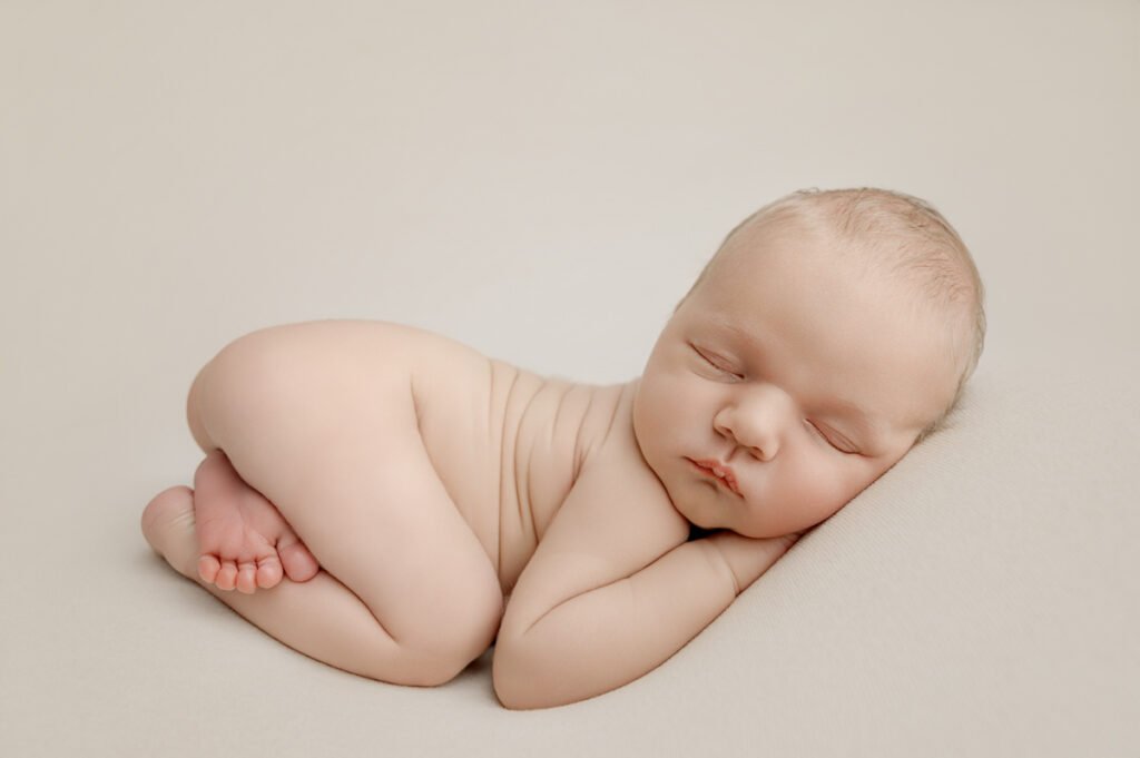 A newborn baby is sleeping on its side on a soft, light-colored surface, with arms and legs gently curled—captured in a serene moment by a skilled newborn photographer.