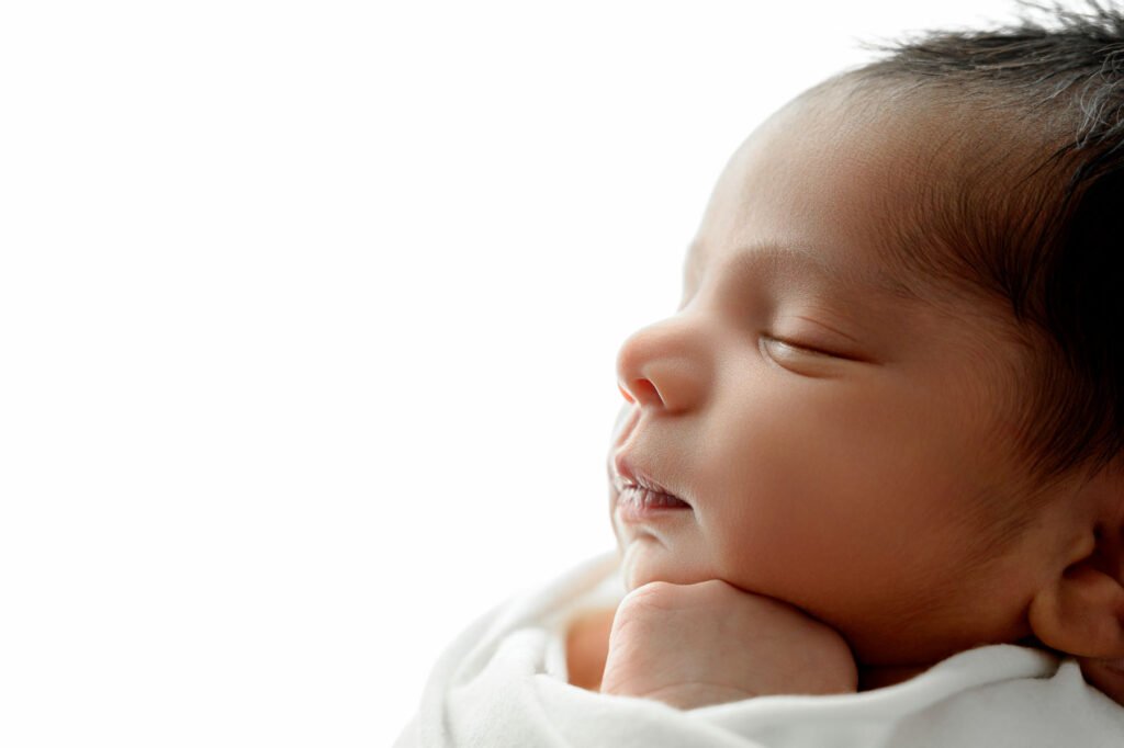 Close-up of a newborn baby sleeping peacefully, wrapped in a white blanket with eyes closed and head resting on hand, against a plain white background. Captured with the gentle artistry of T Haller Photography.
