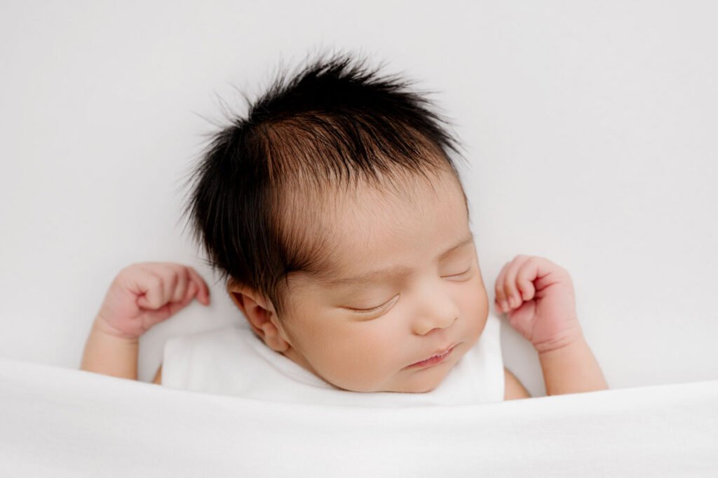 A newborn baby with dark hair sleeps on a white surface, wrapped in a white blanket and wearing a white outfit, beautifully captured by T Haller Photography.