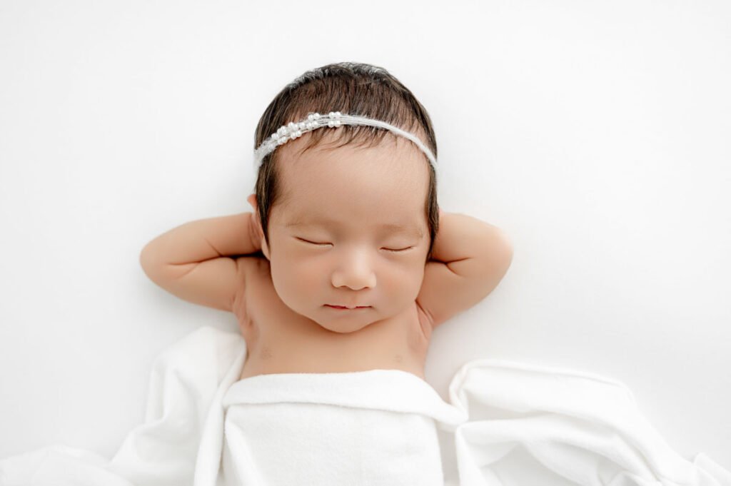 Newborn baby with a white headband lies on a white blanket with eyes closed and arms resting behind head, captured in a serene moment by T Haller Photography.