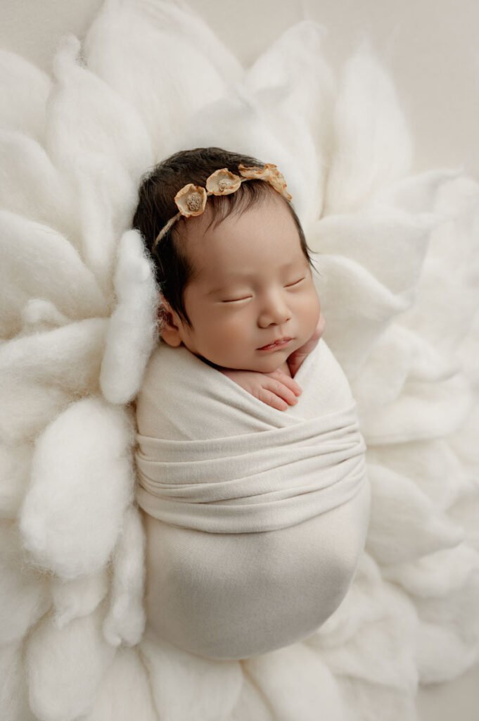 Newborn baby wrapped in a white blanket, lying on a soft, white, flower-shaped surface, wearing a delicate headband with small beige flowers—captured in the timeless style of T Haller Photography.