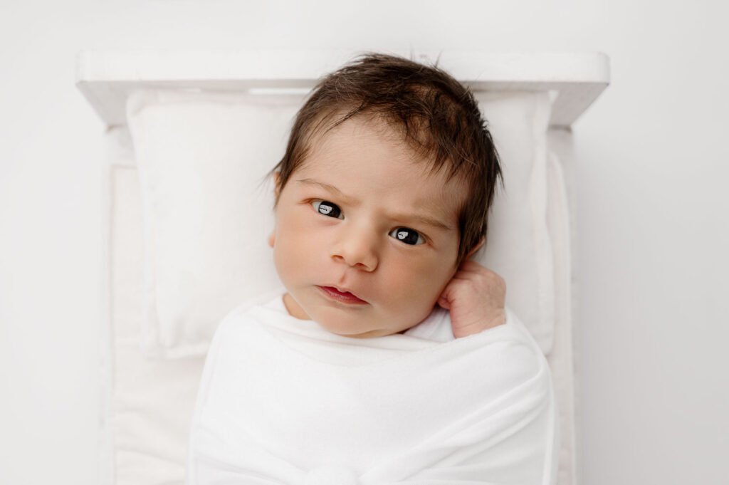 Newborn baby wrapped in a white blanket, lying on a small white bed, looking up with a slightly furrowed brow—beautifully captured by T Haller Photography.