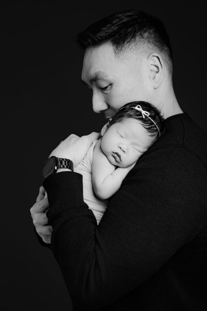 A man holds a sleeping baby with a small bow headband against his chest. Captured by a newborn photographer, the image is in black and white with a dark background.