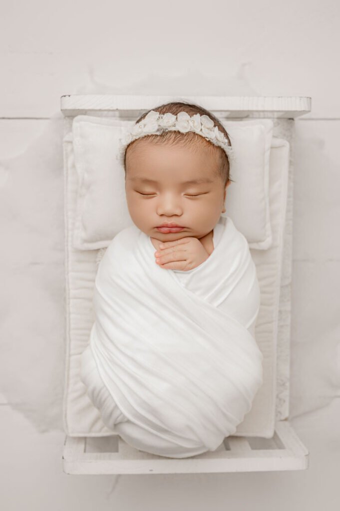 Newborn baby swaddled in a white blanket, wearing a white floral headband, sleeping on a small white bed with white pillows and background—perfectly captured by an experienced newborn photographer.