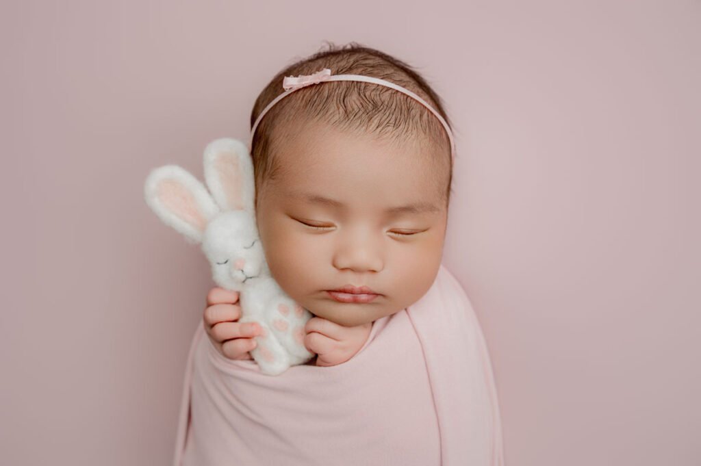 Newborn baby wrapped in a pink blanket, wearing a pink headband, holding a white plush bunny, with eyes closed against a pink background—expertly captured by a skilled newborn photographer.