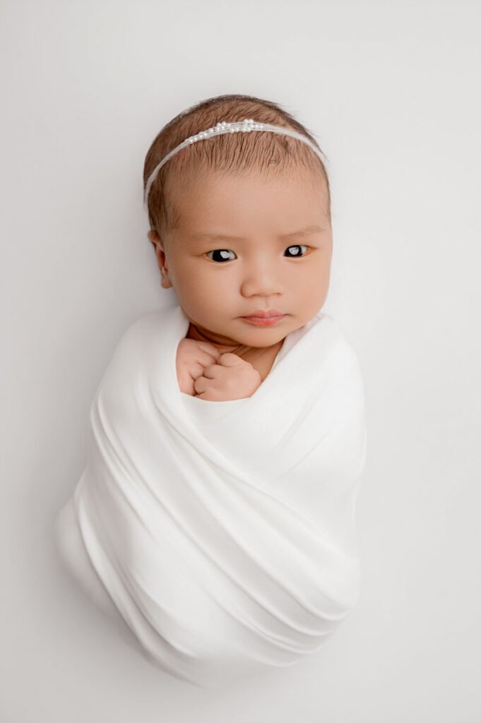 Newborn baby wrapped in a white blanket with a thin headband, lying on a soft white background, looking directly at the camera—a classic portrait captured by a skilled newborn photographer.