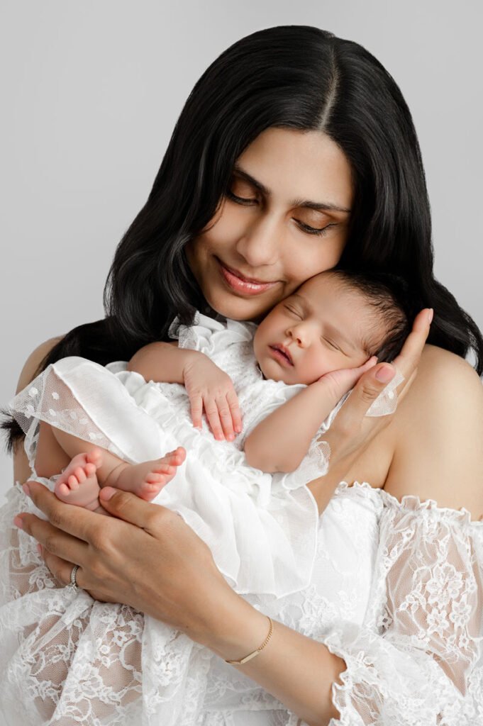 Woman in a white lace dress gently cradles a sleeping baby, both dressed in white, against a plain light grey background—a serene moment beautifully captured by a skilled newborn photographer.